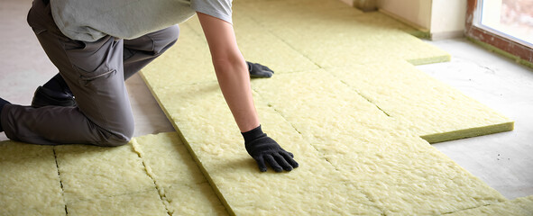 Construction worker installs yellow insulation panels on floor. Man in gray shirt, gray pants, wearing work gloves. Carefully places panels together to create warm floor. Building interior.