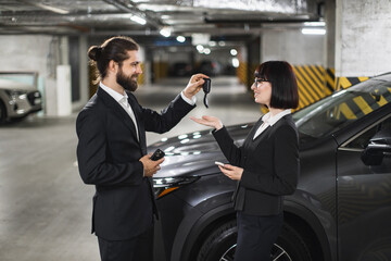 Caucasian businessman giving car keys to Caucasian woman in underground parking lot. Professionals engage in vehicle transaction, emphasizing business, success, and transportation in urban environment
