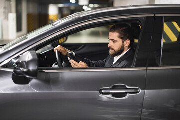 Caucasian male in suit using smartphone inside car parked indoors. Young adult appears focused on device, emphasizing concept of distracted driving.
