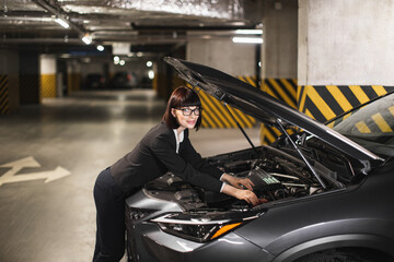 Caucasian businesswoman in formal attire fixes car engine in dimly lit parking garage, conveys...