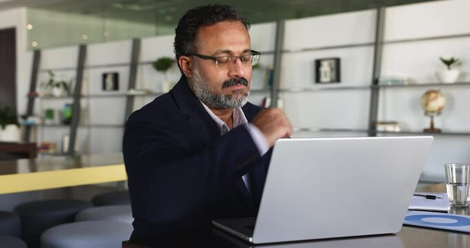 Focused senior Indian business leader man working at laptop computer in co-working office space, touching chin. Executive, company CEO, director man reading online financial report