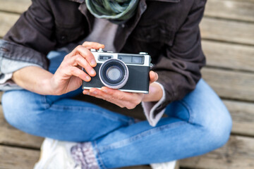 Female photographer sitting cross-legged outdoors on a wooden walkway, holding a vintage camera in her hands, wearing casual clothing, enjoying a moment of creativity and passion for photography
