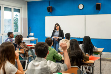 High school teacher giving lesson to diverse group of students raising hands