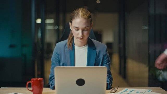 Young woman works on her laptop in a dimly lit office, showing discomfort as a male colleague places a hand on her shoulder. Concept of workplace harassment