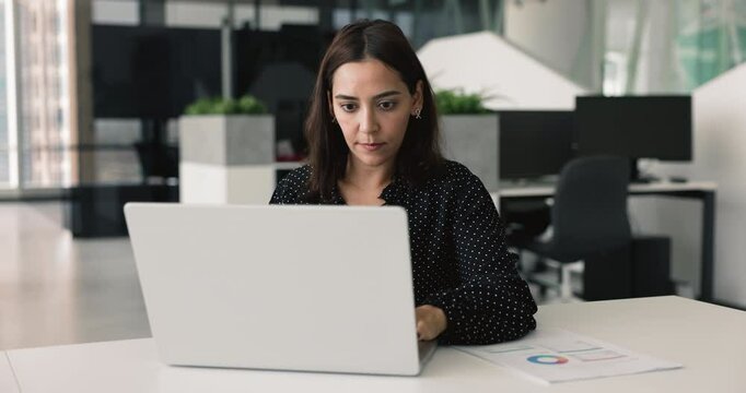 Focused young Middle Eastern business project leader woman working at laptop, in modern office, typing, staring at screen, analyzing marketing statistic research, ecommerce sales on Internet platform
