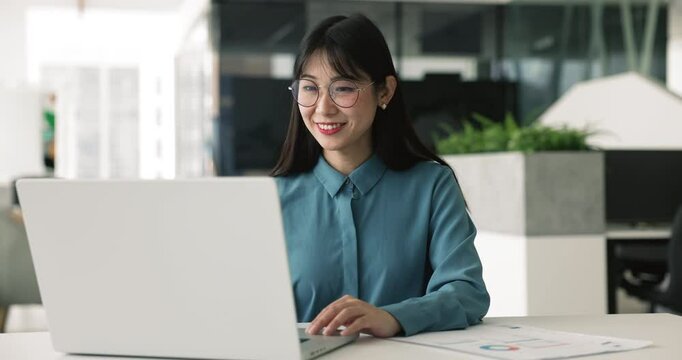 Positive young Asian manager woman in stylish glasses typing on laptop computer at workplace, smiling, using online communication for good job result, analyzing successful marketing business strategy