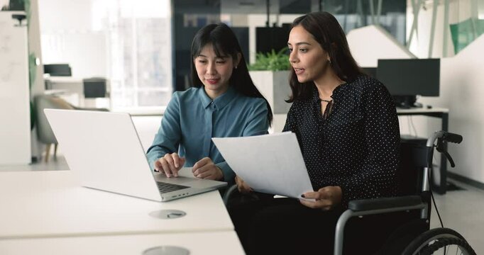 Positive young diverse business colleagues from inclusive company staff working on product together. Young Asian professional woman showing online project content to coworker in wheelchair