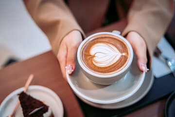 Artistic Latte and Delightful Treat: A pair of elegant hands present a meticulously crafted latte adorned with a heart-shaped design, alongside a tantalizing piece of chocolate cake.