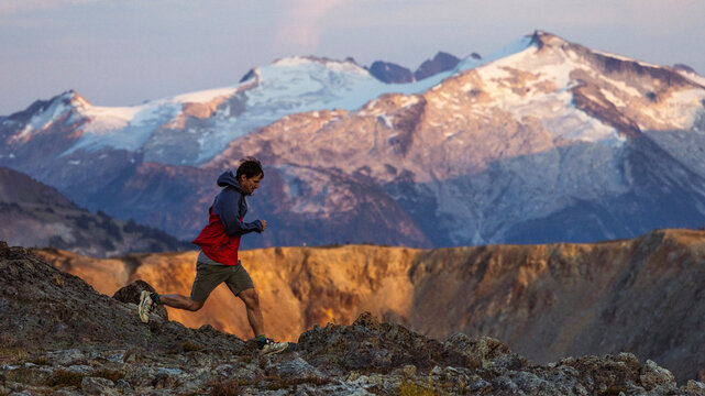 Trail runner leaping across rocks with snow-capped peaks behind.