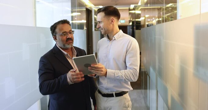 Two diverse different aged male business colleagues discussing online startup, using tablet, standing in office hall. Project manager consulting expert, showing online project on gadget