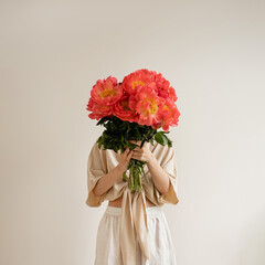 Woman in a white dress presents a lush bouquet of vivid red peonies, set against a simple white...