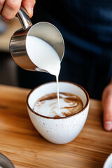 A barista pouring steamed milk into a cup of coffee, creating a creamy swirl. A cozy and inviting coffee moment with a rustic ceramic cup on a wooden surface