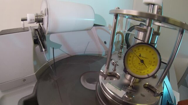 Close-up of a lapping machine with a precision gauge measuring pressure while slurry drips onto the rotating disc for material polishing and refinement