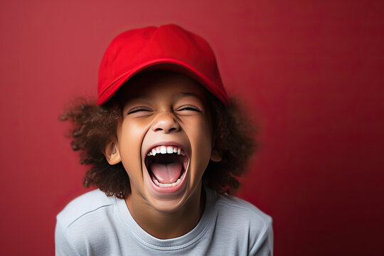 Portrait of a little screaming boy with afro curls in a red cap on a red background.