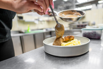 A chef serves a delicious dish featuring meat and mashed potatoes, drizzled with a rich sauce, in a bustling kitchen environment.