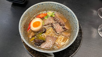 Bowl of beef ramen with egg, beef slices, chili peppers, and sesame seeds. Served in a rustic ceramic bowl. Perfect for illustrating Japanese food, comfort food, and the art of noodle dishes