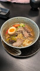 Bowl of beef ramen with egg, beef slices, chili peppers, and sesame seeds. Served in a rustic ceramic bowl. Perfect for illustrating Japanese food, comfort food, and the art of noodle dishes