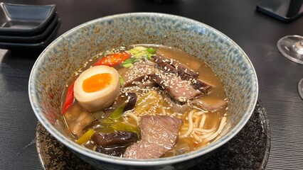Bowl of beef ramen with egg, beef slices, chili peppers, and sesame seeds. Served in a rustic ceramic bowl. Perfect for illustrating Japanese food, comfort food, and the art of noodle dishes