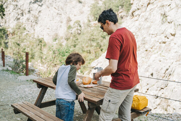 Father dad pours hot coffee tea from thermos into the mug on a family picnic in the mountains. Child school boy kid is watching his dad filling the cup with hot beverage.