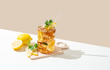Iced tea with lemon, ice and mint in a glass on wooden board on a white and beige background with shadow.