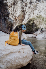 School boy with yellow backpack sits on a riverside rock in the canyon with mountain cliffs in the background. Kid child taking a rest on a boulder near mountain river.