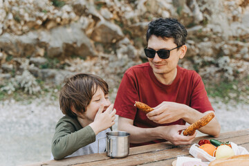 Close-up view of father hugs his school boy son on a family picnic. Child kid and his dad taking a rest and enjoying a picnic while hiking. Boy smiles and bites the bread donut on a picnic.