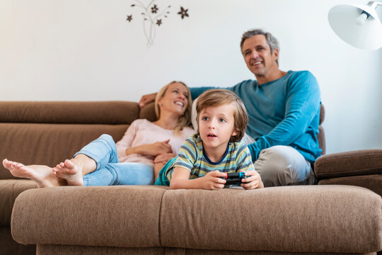 Boy lying on couch at home playing video game with parents watching