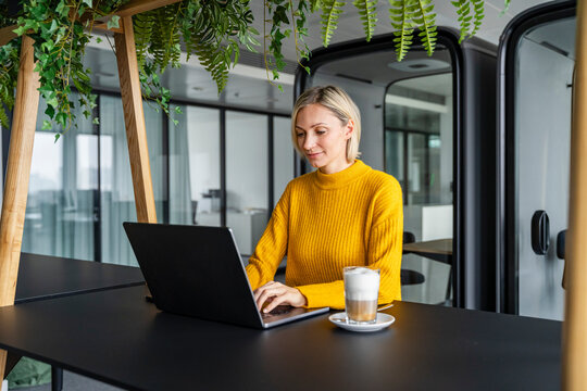Woman working on a laptop in a modern office with greenery and coffee