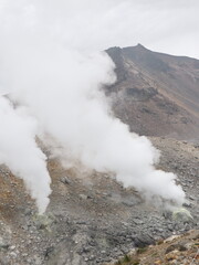 a volcanic landscape with steaming fumaroles or vents emitting gases.  The terrain is rocky and barren, with a mountain slope rising in the background under a cloudy sky.