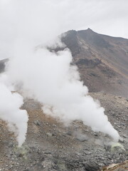 a volcanic landscape with steaming fumaroles or vents emitting gases.  The terrain is rocky and barren, with a mountain slope rising in the background under a cloudy sky.