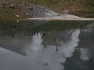 a volcanic landscape with steaming fumaroles or vents emitting gases.  The terrain is rocky and barren, with a mountain slope rising in the background under a cloudy sky.