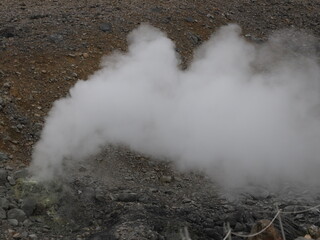 a volcanic landscape with steaming fumaroles or vents emitting gases.  The terrain is rocky and...