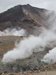 a volcanic landscape with steaming fumaroles or vents emitting gases.  The terrain is rocky and barren, with a mountain slope rising in the background under a cloudy sky.