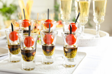 Tasty canapes with cherry tomatoes, mozzarella, balsamic vinegar and pesto sauce in shot glasses on white table, closeup