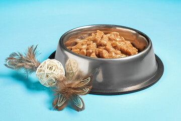 Wet pet food in feeding bowl and cat toy on light blue background