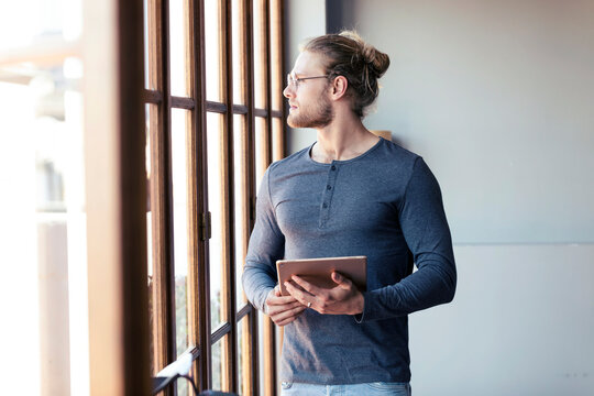 Pensive young man at home with a digital tablet looking out of a window