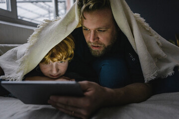 Father and son lying together under blanket looking at digital tablet