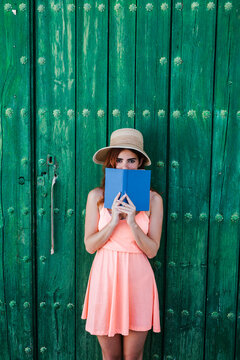 Portrait of redheaded young woman standing in front of green wooden door covering mouth with a book