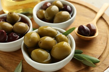 Delicious marinated olives in bowls, spoon and green leaves on table, closeup