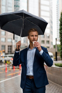 Young businessman with umbrella drinking a coffee during a rainy day in Bangkok