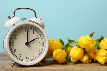 Spring time. Alarm clock and beautiful tulips on wooden table against light blue background, closeup