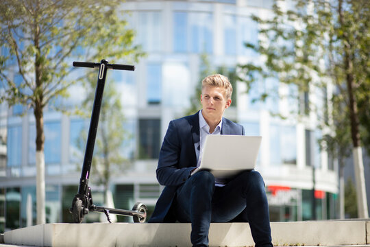 Young businessman with e-scooter using laptop in the city