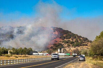 Wildfire smoke blankets hillside, traffic pauses. People observe the blaze from roadside. ,...