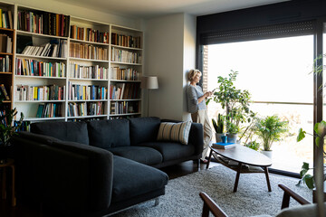 Mature woman standing at the window in living room at home using smartphone