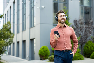 A handsome man walks outside near an office building, enjoying music on his headphones while using...