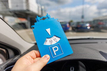 Driver hand hold blue parking disc show arrival time near large shopping complex mall.  Parking time restrictions in busy parking lots. Limited time duration park place zone