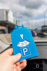 Driver hand hold blue parking disc show arrival time near large shopping complex mall.  Parking...