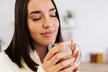 Young pretty woman in housecoat smelling the aroma of coffee in the morning standing in her kitchen...