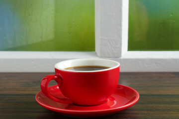 Red cup of aromatic coffee and saucer on wooden sill near window, closeup
