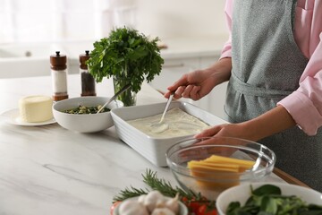 Woman spreading bechamel sauce onto spinach lasagna at marble table indoors, closeup
